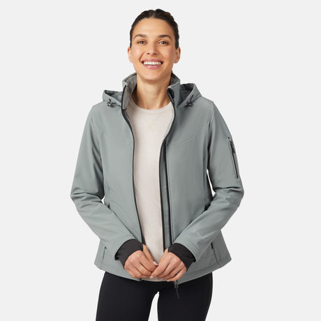 A smiling woman wears the Free Country Women's StormTech Super Softshell® Jacket in light gray over a white top and black pants, standing against a plain white background.