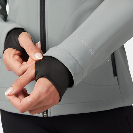 A woman wears the Free Country Women’s StormTech Super Softshell® Jacket in light gray with black thumbhole cuffs and a zipper, adjusting the sleeve where “FREE COUNTRY” is printed—highlighting quality outdoor apparel. Only torso and hands are shown.