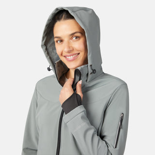 A woman smiles at the camera in the Free Country Women's StormTech Super Softshell® Jacket in light gray, holding the zipper near her chin. The water-resistant jacket has a sleeve zip pocket and cuff thumb holes, shown against a plain white background.