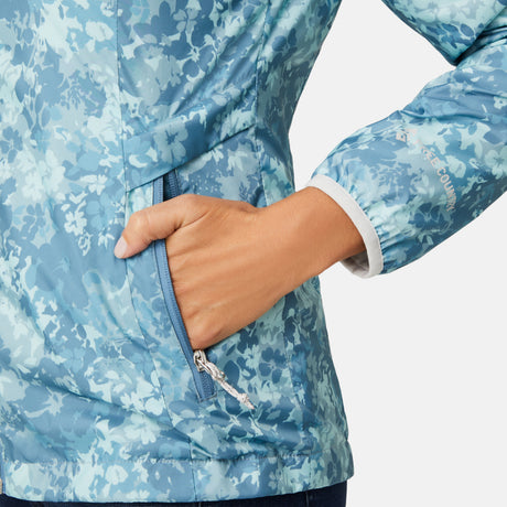 A person rests their right hand in the zippered pocket of the Clearance Women's Windshear Jacket—a blue, floral-patterned, water-resistant windbreaker with white cuff lining and a partially zipped pocket.