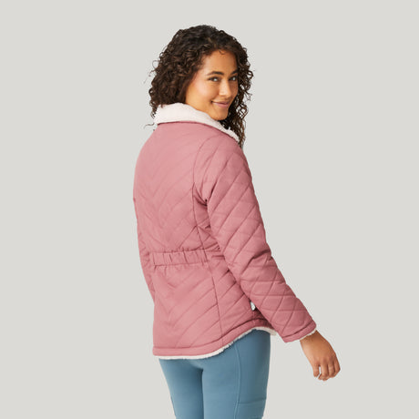 A woman with curly hair, wearing a Clearance Women's FreeCycle® Cloud Lite II Reversible Jacket in light pink with a white sherpa collar and blue pants, stands facing away, smiling over her shoulder against a plain light background.
