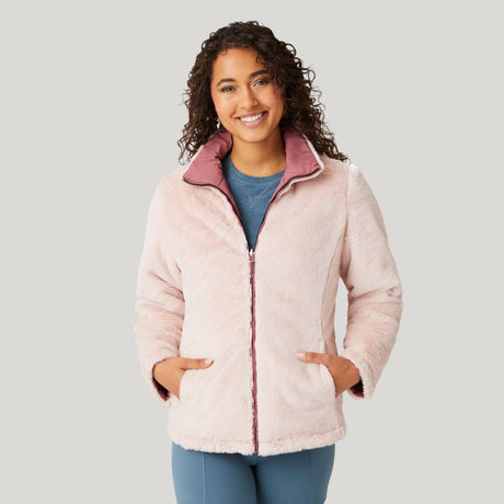 A smiling woman with curly hair wears the Women's FreeCycle® Cloud Lite II Reversible Jacket by Clearance in light pink with a mauve collar, over a blue shirt and light blue pants, standing against a plain light background.