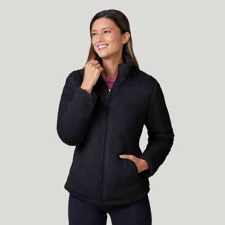 A woman smiles with one hand in her pocket and the other touching the collar of a wind-resistant black Women's FreeCycle® Cloud Lite II Reversible Jacket by Clearance, standing against a light gray background.