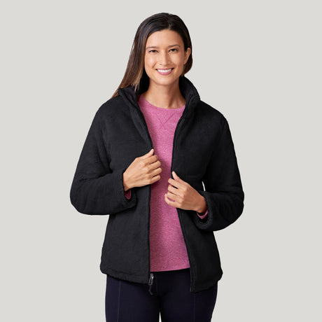 A woman with long brown hair smiles wearing the Clearance Women's FreeCycle® Cloud Lite II Reversible Jacket, black and wind resistant, over a pink shirt and dark pants against a plain light gray background.