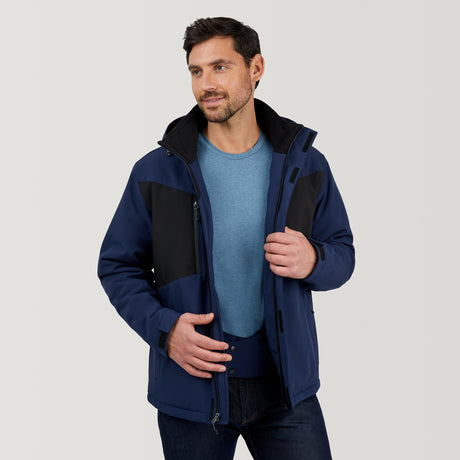 A man in a Clearance Men's Thermo Softshell® Mid Weight Jacket, blue and black and water resistant, stands against a plain light background, looking to the side with a slight smile and holding his jacket front with one hand.