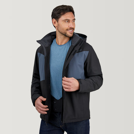 A man in a Clearance Men's Thermo Softshell® Mid Weight Jacket (black and gray) over a blue shirt stands against a light background, smiling and looking left while adjusting his jacket.