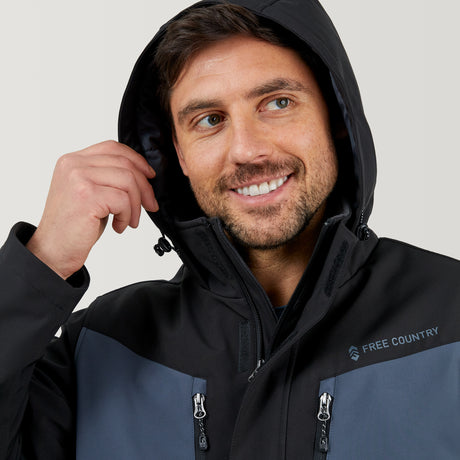 A bearded man smiles while wearing the Men's Thermo Softshell® Mid Weight Jacket by Clearance, featuring a black and gray water-resistant design. He holds the hood with one hand, and "FREE COUNTRY" is displayed on the chest.