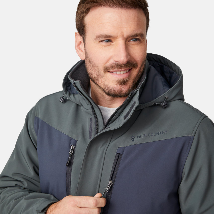 [Joe is 6'0" wearing size M.] Men’s Free Country two-tone hooded zip-front jacket with dark gray body and black paneling across shoulders and chest—features dual vertical zippered chest pockets, side hand pockets, full-length front zipper, and an attached interior snow skirt for enhanced protection against the elements. Paired with matching gray pants for a sleek, coordinated winter look.