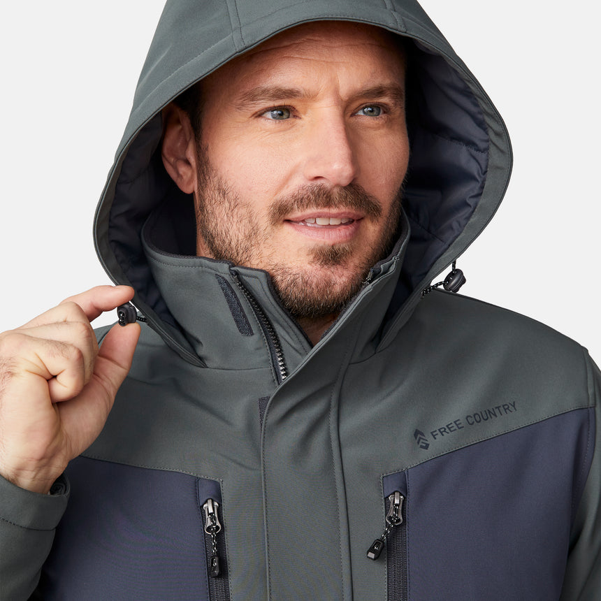 [Joe is 6'0" wearing size M.] Men’s Free Country two-tone hooded zip-front jacket with dark gray body and black paneling across shoulders and chest—features dual vertical zippered chest pockets, side hand pockets, full-length front zipper, and an attached interior snow skirt for enhanced protection against the elements. Paired with matching gray pants for a sleek, coordinated winter look.