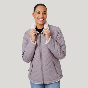 A woman smiles while wearing the Clearance Women's FreeCycle® Cloud Lite II Reversible Jacket, a light purple quilted style with a white fleece collar made from recycled plastic bottles, paired with blue jeans and a black top against a gray background.