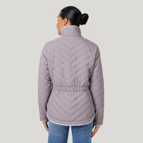 A woman with dark hair in a bun wears the Clearance Women's FreeCycle® Cloud Lite II Reversible Jacket in light purple and blue jeans, shown from the back against a plain light gray background.