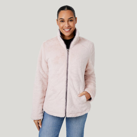 A woman smiles with her left hand in the pocket of the Women's FreeCycle® Cloud Lite II Reversible Jacket by Clearance—a light pink, plush faux fur jacket made from recycled plastic bottles. She wears blue jeans and stands against a light gray background.
