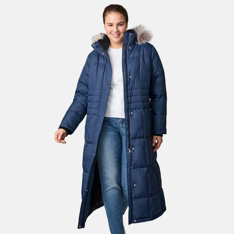 A woman smiles and walks forward in a Free Country Women's Plus Size Full Length Splendor Down Jacket with a fur-lined hood, paired with a white top and blue jeans, against a plain light gray background.