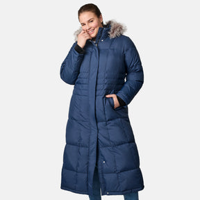 A smiling woman wears the Free Country Women's Plus Size Full Length Splendor Down Jacket, featuring a blue water-resistant finish and faux fur-lined hood, posing with one hand on her face and the other in her pocket against a plain background.