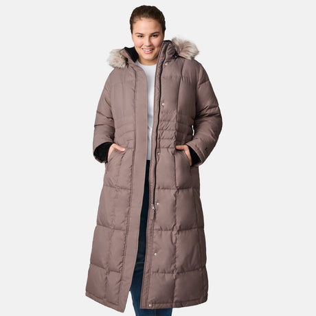A woman smiles at the camera with her hands in her pockets, wearing the Free Country Women's Plus Size Full Length Splendor Down Jacket. The taupe parka features a faux fur-lined hood and water-resistant warmth against a plain white background.