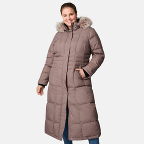 A woman smiles with one hand in her pocket, wearing the Free Country Women's Plus Size Full Length Splendor Down Jacket in taupe with a faux fur–trimmed hood and water-resistant finish, set against a plain white background.