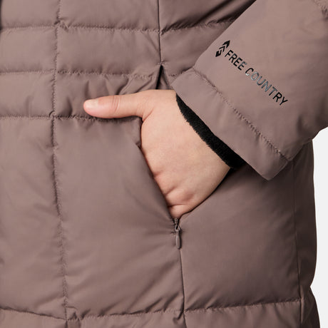 A woman wears the Free Country Women's Plus Size Full Length Splendor Down Jacket in taupe, featuring water-resistant quilted fabric, zippered side pockets, and the brand's black logo on the sleeve.
