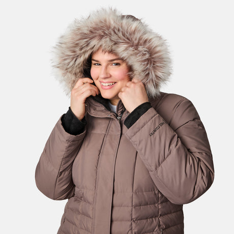 A woman smiles while wearing the Free Country Women's Plus Size Full Length Splendor Down Jacket, a water-resistant brown winter coat featuring a faux fur-lined hood, holding the hood with both hands against a white background.