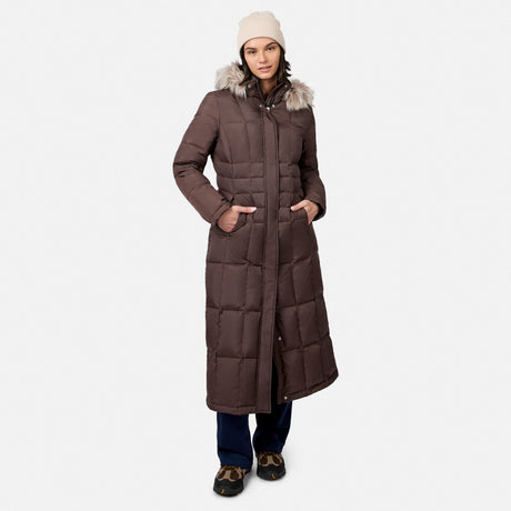 [Model is wearing size S.] A woman modeling a long brown puffer parka with a faux-fur trimmed hood, paired with a cream beanie, dark pants, and brown shoes, against a light gray studio background.