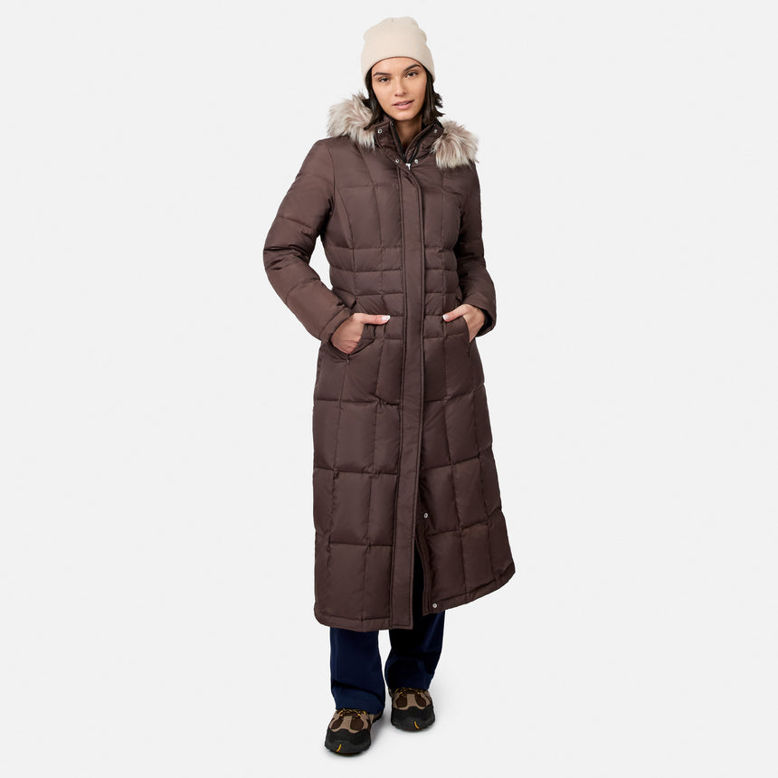 [Model is wearing size S.] A woman modeling a long brown puffer parka with a faux-fur trimmed hood, paired with a cream beanie, dark pants, and brown shoes, against a light gray studio background.