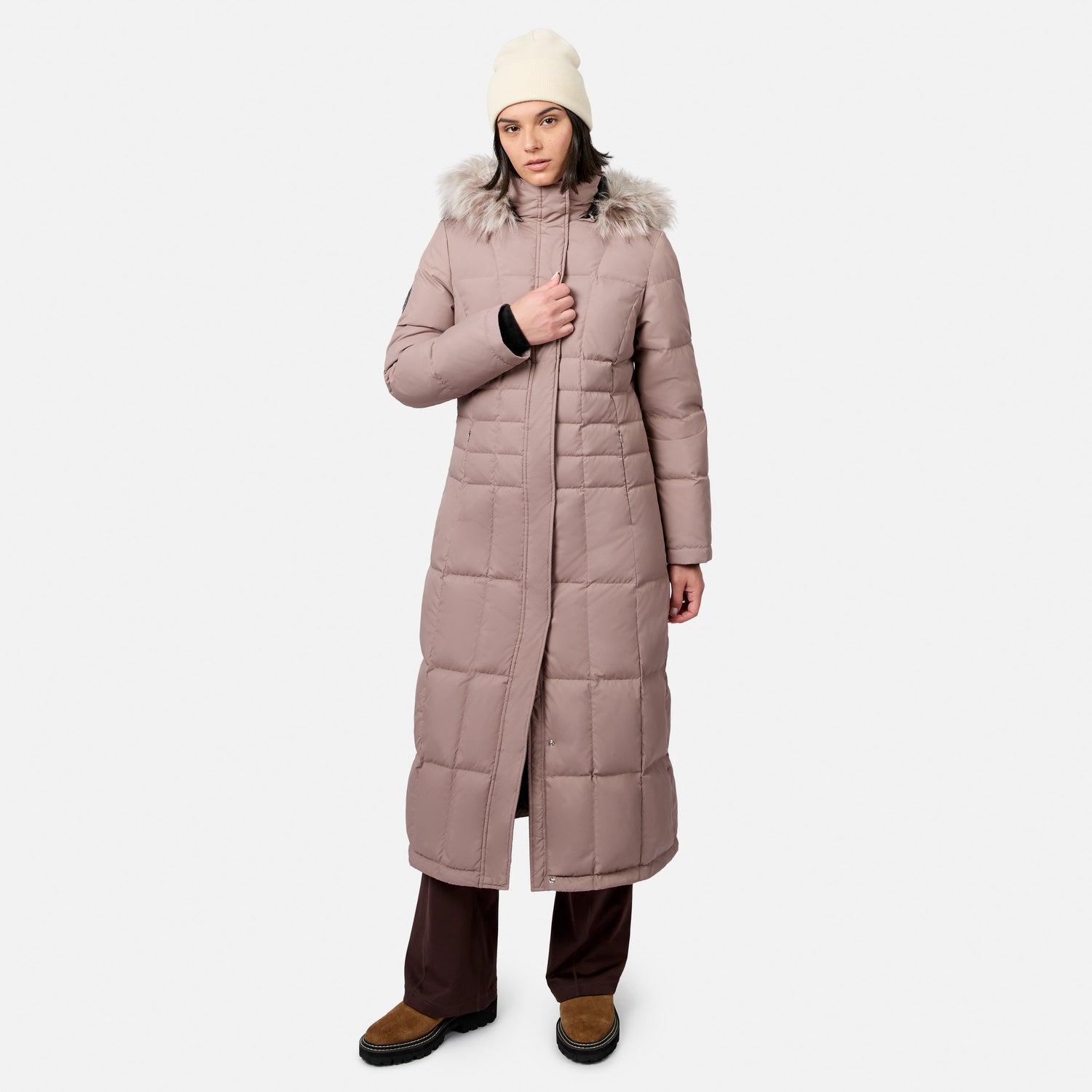 Woman wearing a full length dusty rose Splendor Down Jacket with a fur trimmed hood and cream knit beanie