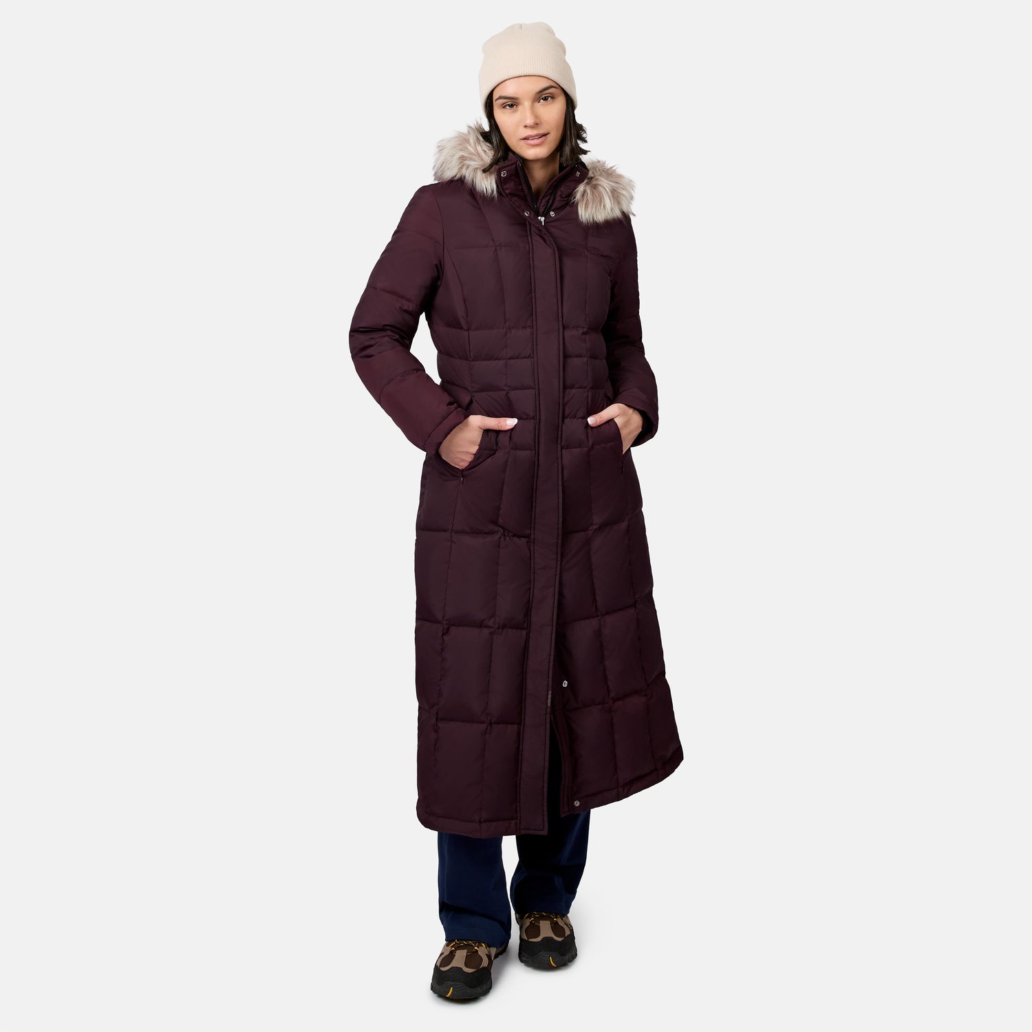 Front view of a woman in a burgundy full length Splendor down jacket with fur trimmed hood wearing a beige beanie