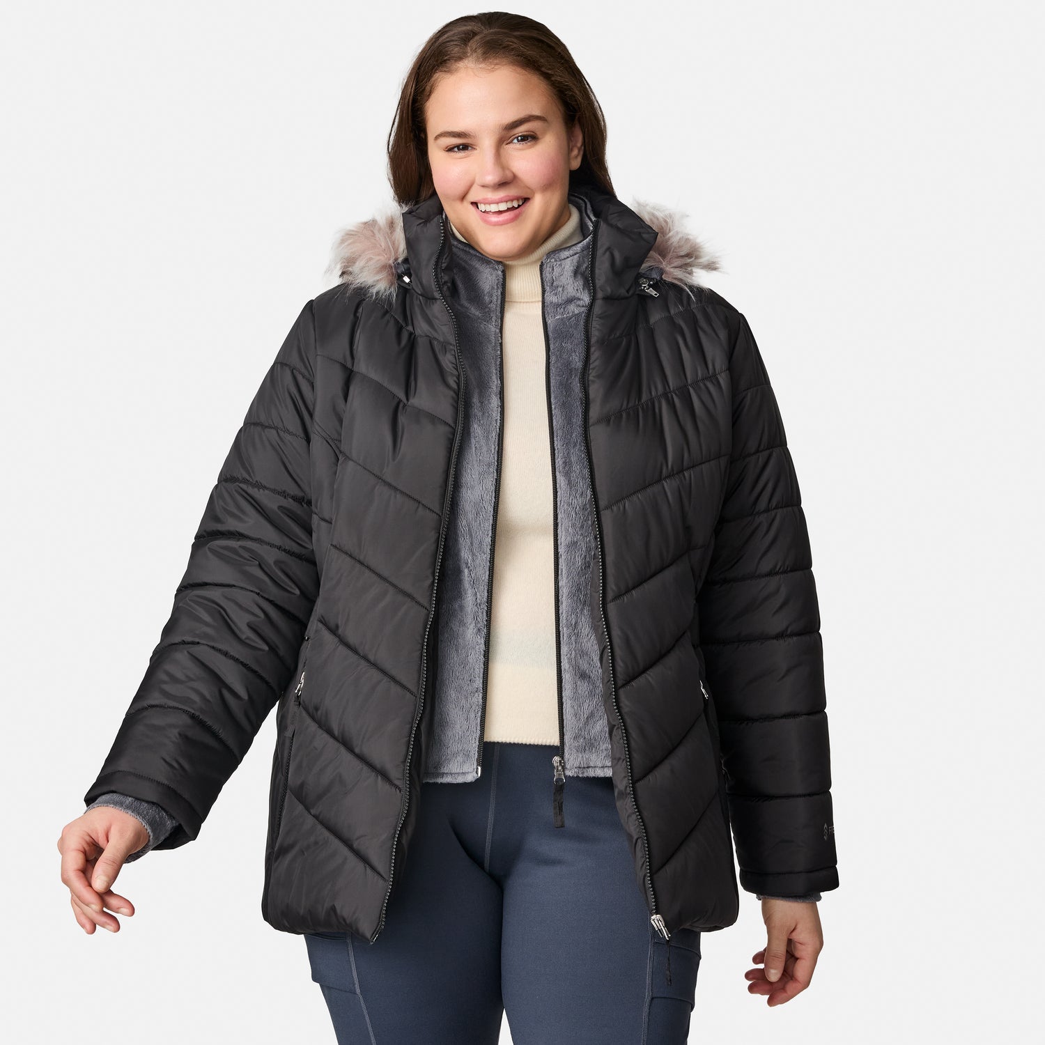 A woman smiles while wearing the Free Country Women’s Plus Size Unstoppable II Poly Air Touch Jacket in black, featuring a faux fur-trimmed hood and eco-friendly REPREVE fill, paired with a cream sweater and navy pants against a plain light gray background.