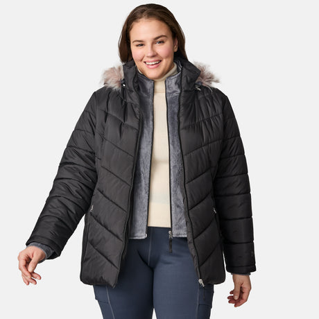 A woman smiles while wearing the Free Country Women’s Plus Size Unstoppable II Poly Air Touch Jacket in black, featuring a faux fur-trimmed hood and eco-friendly REPREVE fill, paired with a cream sweater and navy pants against a plain light gray background.