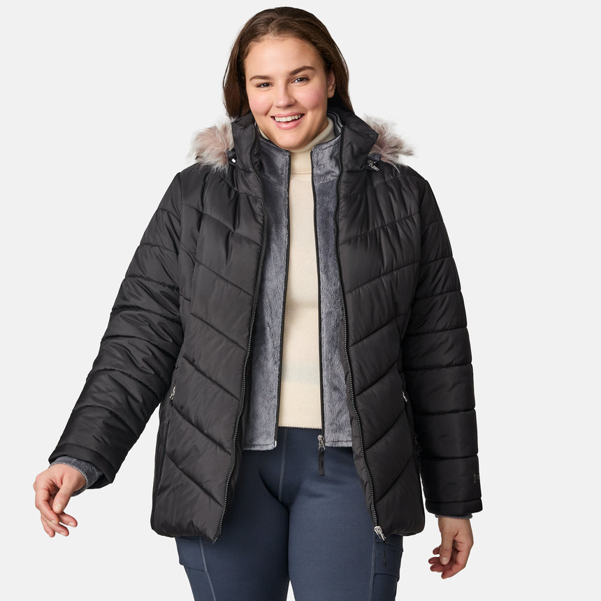 A woman smiles while wearing the Free Country Women’s Plus Size Unstoppable II Poly Air Touch Jacket in black, featuring a faux fur-trimmed hood and eco-friendly REPREVE fill, paired with a cream sweater and navy pants against a plain light gray background.