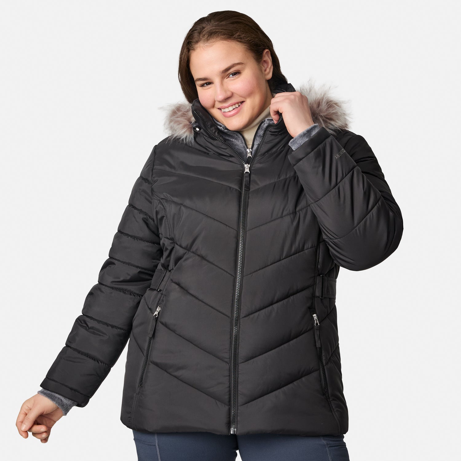 A woman is smiling and posing with one hand touching the hood while wearing the Free Country Women’s Plus Size Unstoppable II Poly Air Touch Jacket in black. This jacket features lightweight eco-friendly REPREVE insulation and a faux fur-trimmed hood, offering both warmth and style.