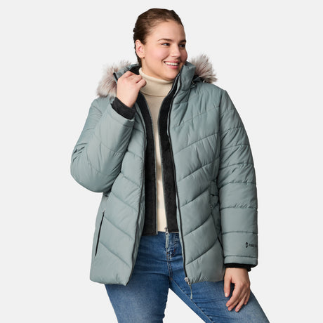 A smiling woman models the Free Country Women's Plus Size Unstoppable II Poly Air Touch Jacket, a light blue quilted winter coat with a faux fur-trimmed hood, over a cream turtleneck and blue jeans against a white background.