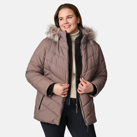 A smiling woman wears the Free Country Women's Plus Size Unstoppable II Poly Air Touch Jacket in mauve with REPREVE fill and a faux fur-trimmed hood, layered over a cream turtleneck and black pants against a plain white background.