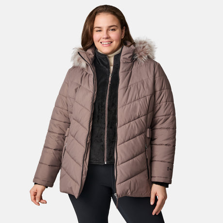 A smiling woman wears the Free Country Women's Plus Size Unstoppable II Poly Air Touch Jacket, a mauve quilted eco-friendly winter coat with REPREVE fill and faux fur-trimmed hood, over a black zip-up and pants, against a light gray background.
