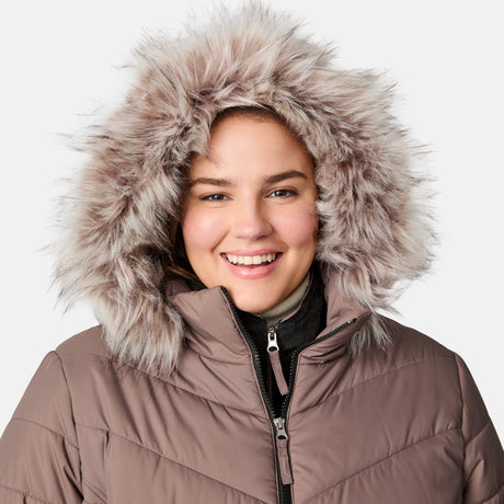 A smiling woman stands against a light background wearing the Free Country Women's Plus Size Unstoppable II Poly Air Touch Jacket, featuring an eco-friendly taupe winter design and a large, fluffy faux fur hood.
