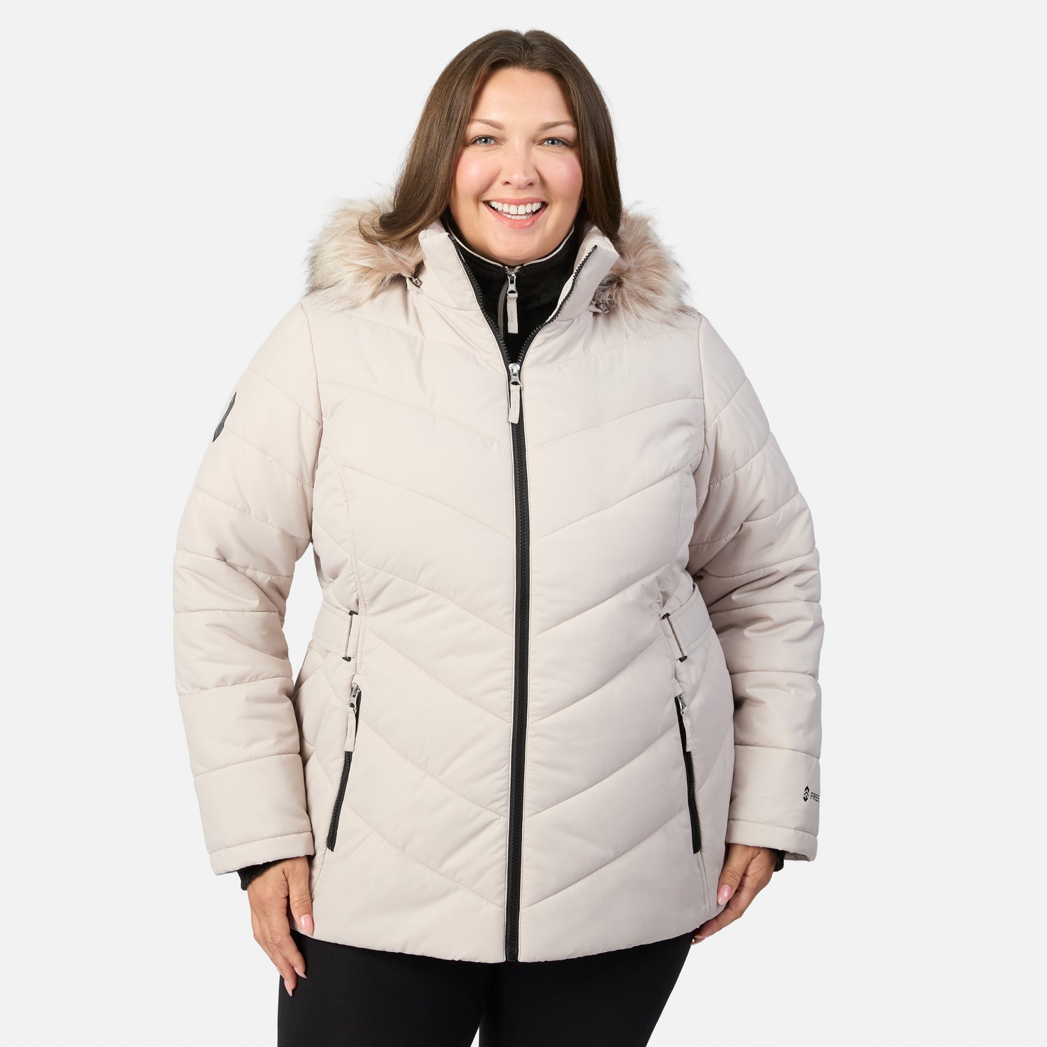 [Model is wearing size 1X] A woman wearing a light beige quilted winter parka with a faux-fur trimmed hood, black zipper details, and black pants, photographed against a plain light gray studio background.