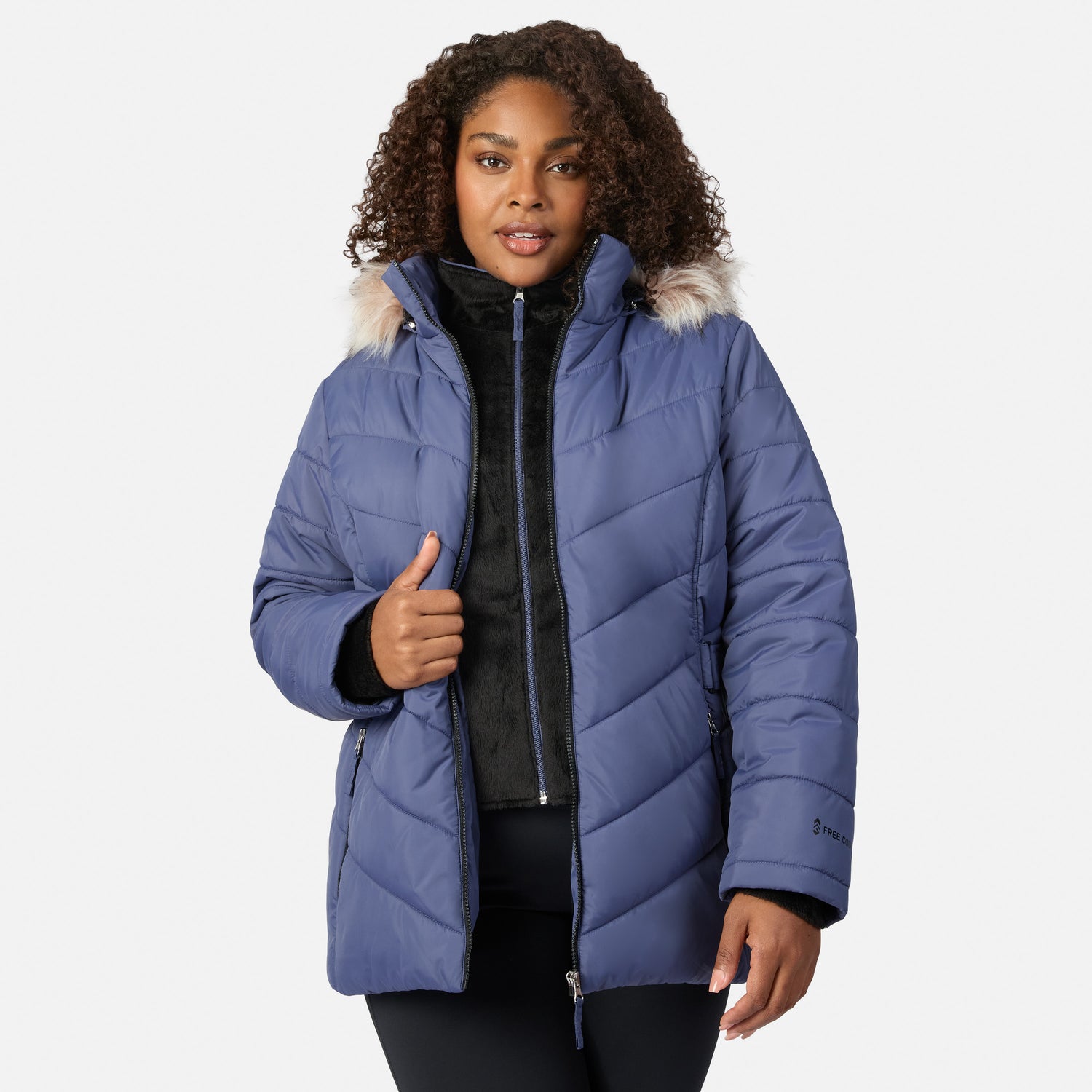 [Model is wearing size 1X] A woman wearing a blue quilted puffer jacket with a light beige faux-fur trimmed hood, layered over a black zip-up fleece. She is standing against a solid light gray background, giving a thumbs-up with her right hand.