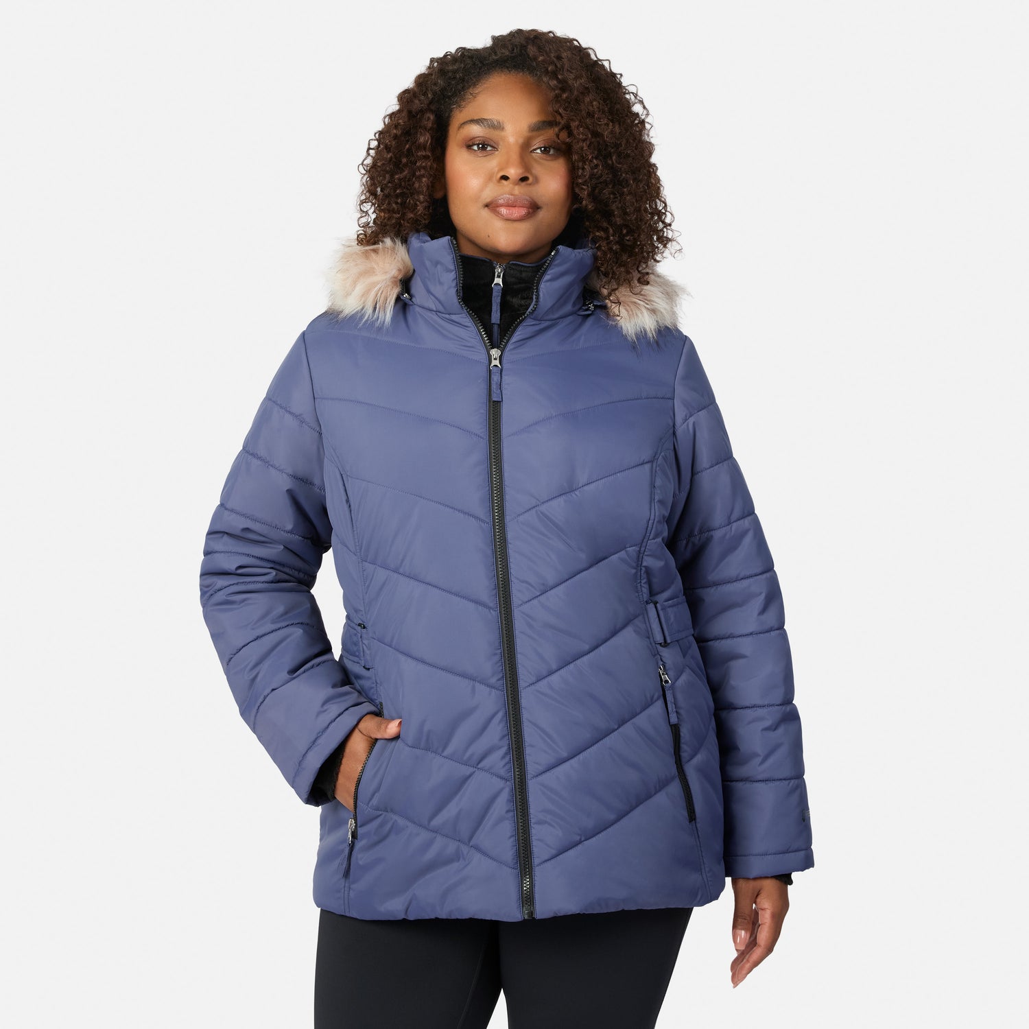 [Model is wearing size 1X] A woman wearing a slate blue quilted puffer jacket with a faux-fur trimmed hood and black zipper details, photographed against a light gray studio background.