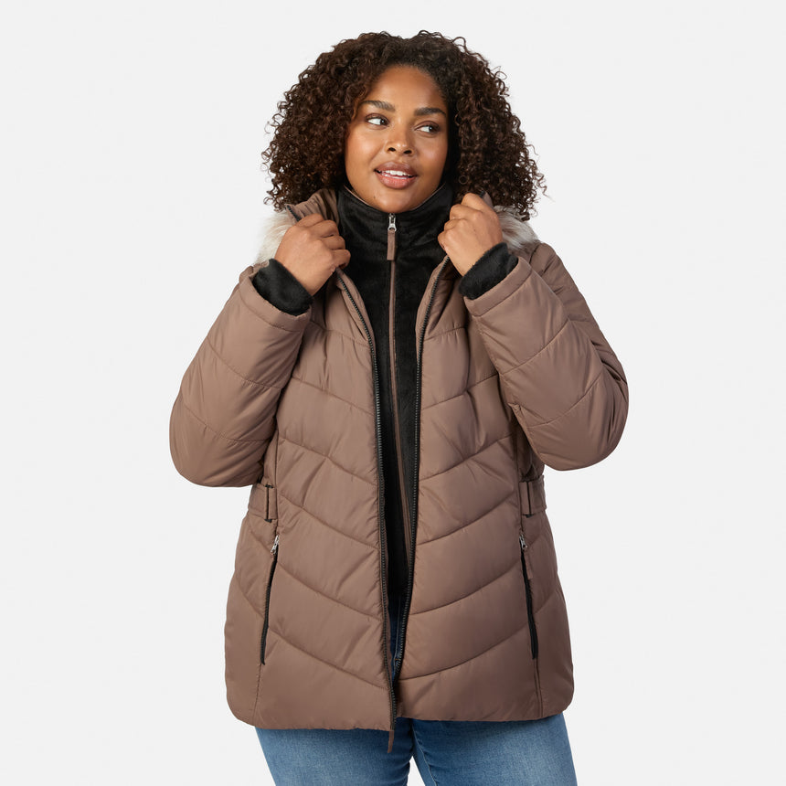 [Model is wearing size 1X] A woman with curly brown hair wearing a taupe-brown puffer jacket with a black inner collar and zipper, styled over a black top and blue jeans, against a light gray studio background.