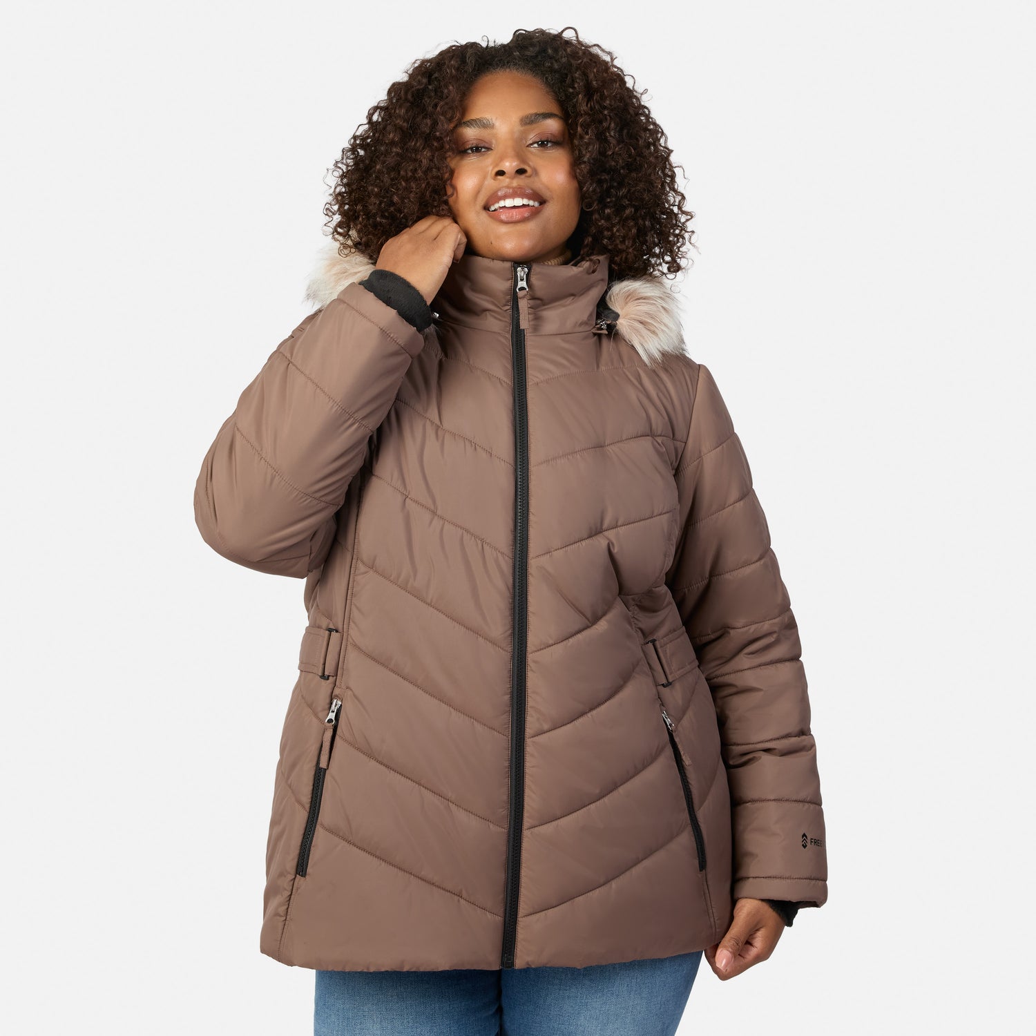 [Model is wearing size 1X] A woman wearing a taupe-brown quilted puffer jacket with a light beige faux-fur trimmed hood and black zippers, paired with blue jeans, against a light gray studio background.