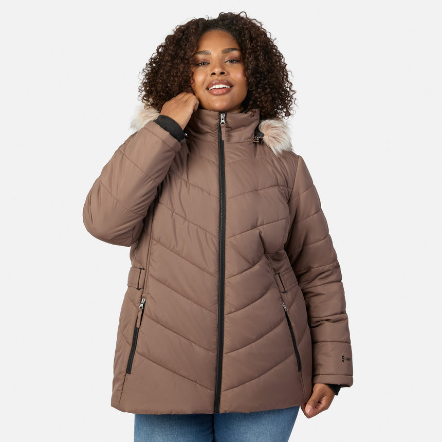 [Model is wearing size 1X] A woman wearing a taupe-brown quilted puffer jacket with a light beige faux-fur trimmed hood and black zippers, paired with blue jeans, against a light gray studio background.