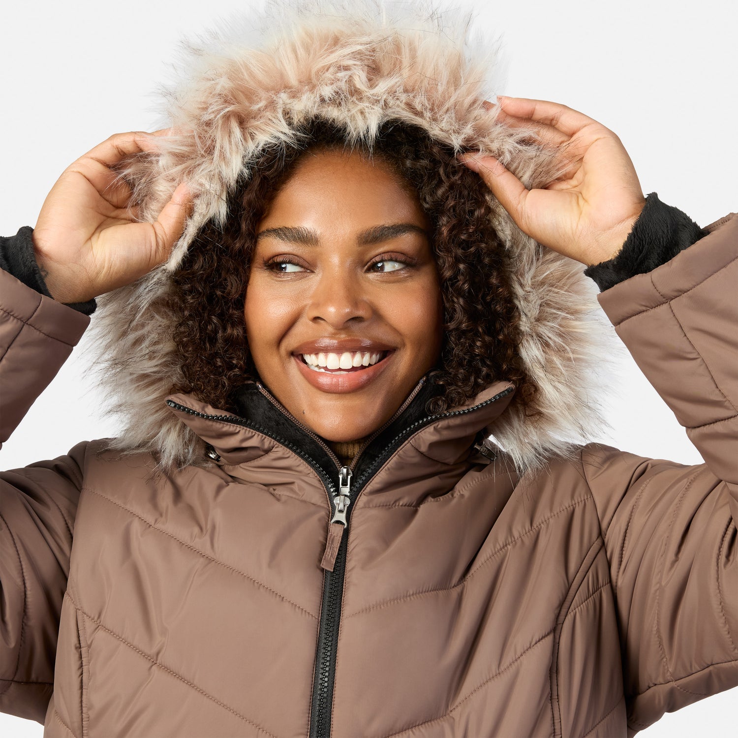 [Model is wearing size 1X] A smiling woman wearing a brown quilted puffer jacket with a large fluffy beige faux-fur trimmed hood, adjusting the hood with both hands against a light gray background.
