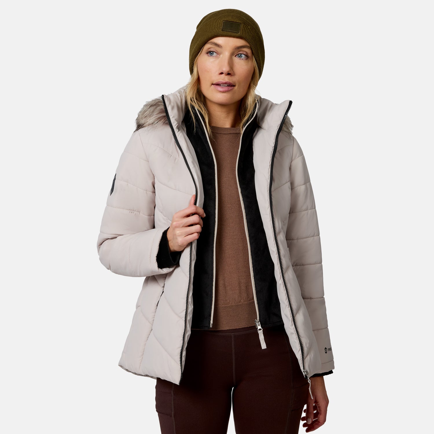 [Model is wearing size S.] A woman wearing a light beige puffer jacket with black trim, a brown sweater, dark brown pants, and an olive green beanie, shown against a plain light gray studio background.