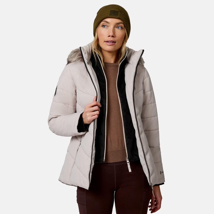 [Model is wearing size S.] A woman wearing a light beige puffer jacket with black trim, a brown sweater, dark brown pants, and an olive green beanie, shown against a plain light gray studio background.