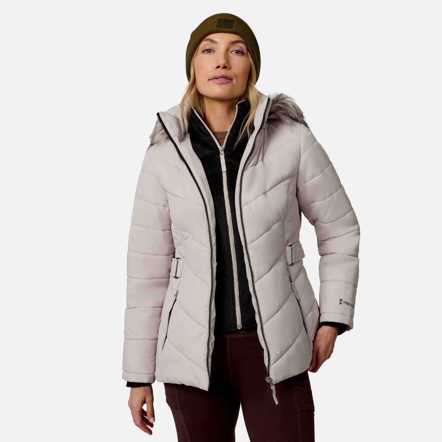 [Model is wearing size S.] A woman wearing a light beige hooded puffer jacket with a faux-fur trimmed hood, a black zip-up fleece layer, brown pants, and an olive green beanie, photographed against a light gray studio background.