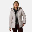 [Model is wearing size S.] A woman wearing a light beige hooded puffer jacket with a faux-fur trimmed hood, a black zip-up fleece layer, brown pants, and an olive green beanie, photographed against a light gray studio background.