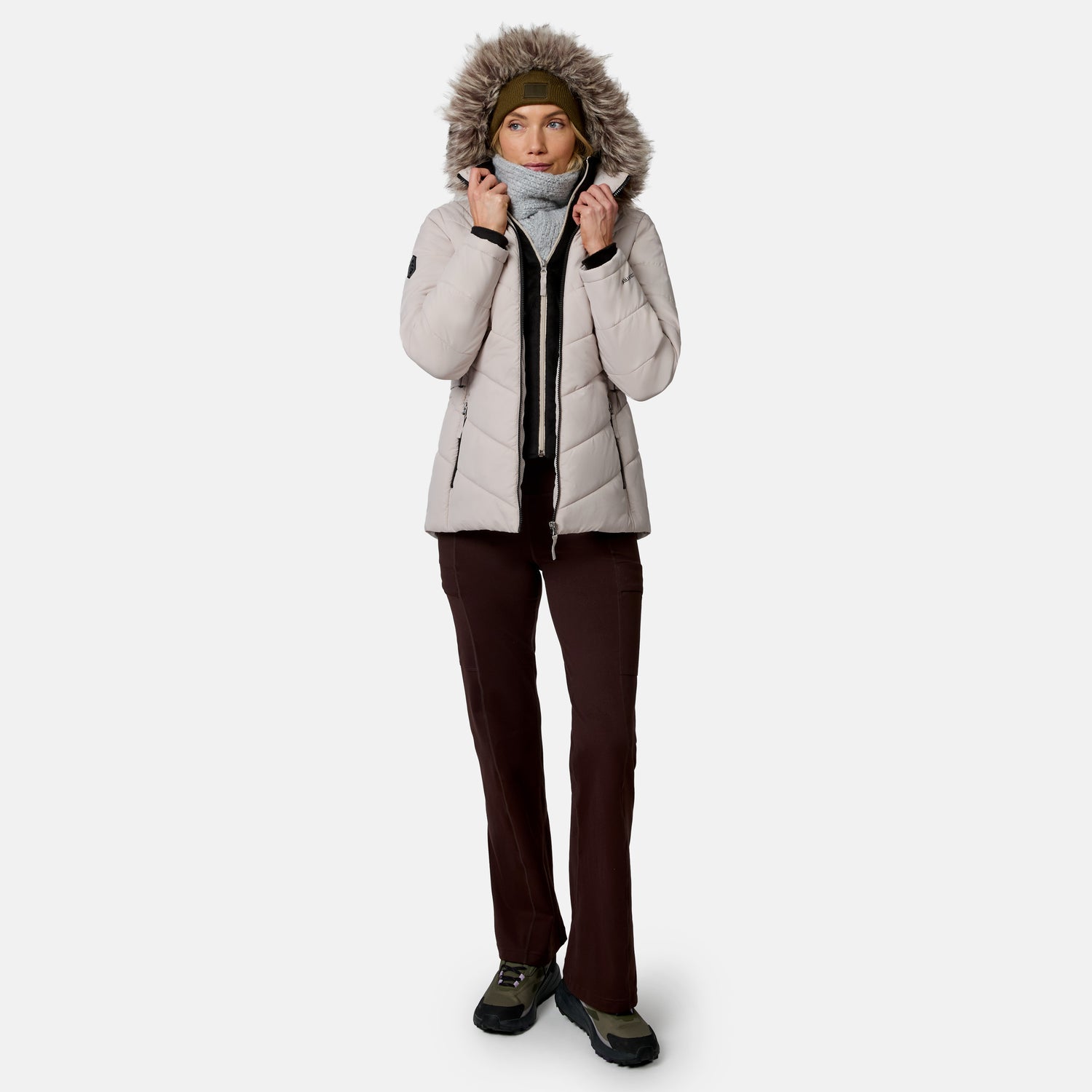 [Model is wearing size S.] A woman modeling a light beige hooded puffer jacket with a faux-fur trimmed hood, dark brown pants, and hiking boots against a plain light gray studio background.
