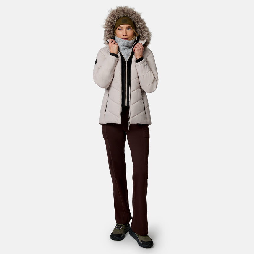 [Model is wearing size S.] A woman modeling a light beige hooded puffer jacket with a faux-fur trimmed hood, dark brown pants, and hiking boots against a plain light gray studio background.