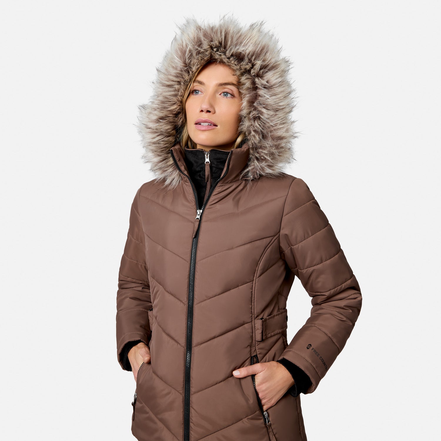 [Model is wearing size S.] A woman wearing a brown quilted winter parka with a large faux-fur trimmed hood, standing against a light gray studio background.