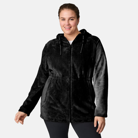 A woman wears a black Free Country Women’s Plus Size Grid Butter Pile® Anorak Jacket and black leggings, standing against a light background, smiling with one hand gently raised.