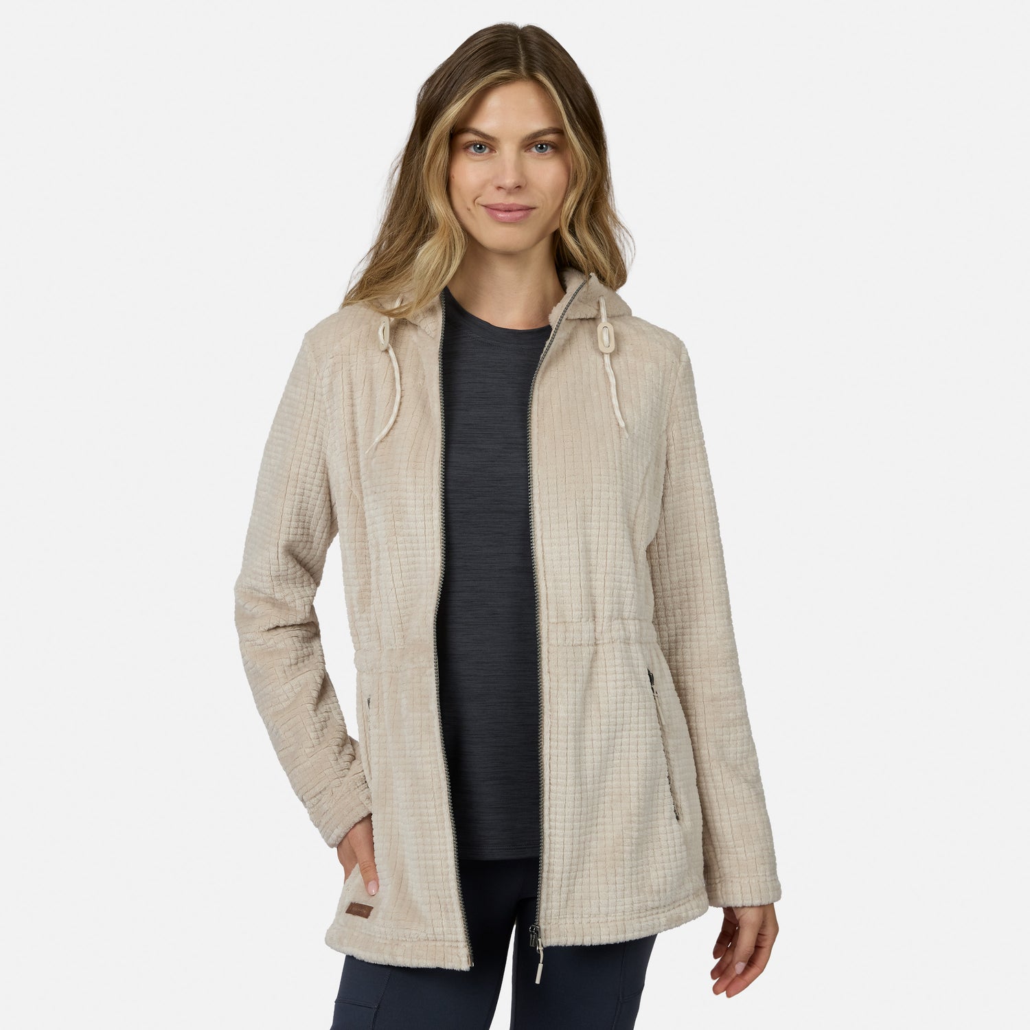 A woman models the Free Country Women’s Grid Butter Pile® Anorak Jacket in light beige, hooded with drawstrings, over a dark shirt and pants, standing with one hand in her pocket against a plain white background.